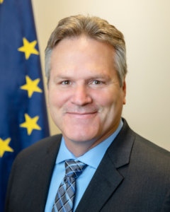 Alaska Governor Mike Dunleavy smiling in front of the Alaska state flag