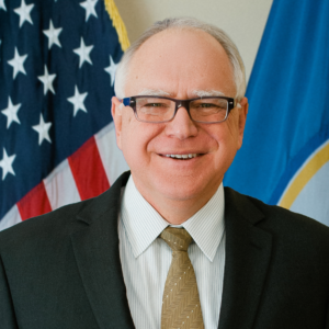 Governor Tim Walz smiling in front of an American flag and the Minnesota state flag