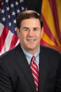Governor Doug Ducey smiling in front of an American flag and the Arizona state flag