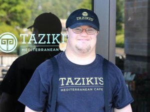 A worker with a disability standing outside Taziki's Mediterranean Cafe wearing a hat and shirt with the restaurant's logo on it