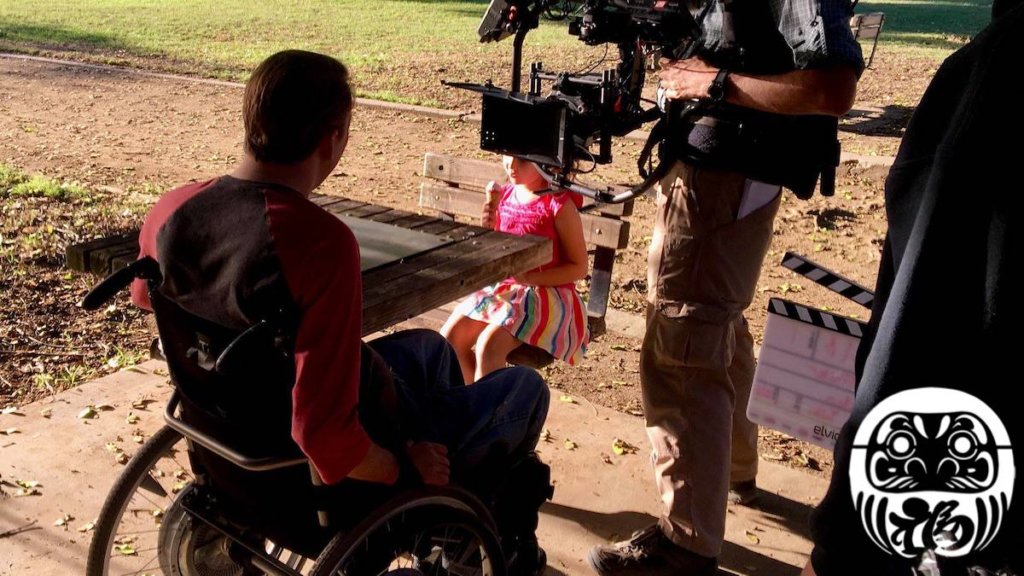 Tobias Forrest and a young girl on the set of Daruma with a camera pointed at Tobias. Logo for Daruma in bottom right.