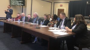Eight panelists sitting behind a table in a Capitol Hill meeting room for an NDEAM event. Sign language interpreter and screen with CART text on left side of photo.