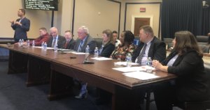 Eight panelists sitting behind a table in a Capitol Hill meeting room for an NDEAM event. Sign language interpreter and screen with CART text on left side of photo.