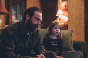 Aaron Paul and a young girl sitting on a couch inside of a living room setting