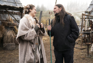 A scene from See with two actors outside smiling at each other in front of grass, trees and huts.