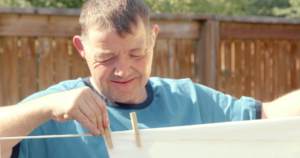 A man with a disability hanging up clothes on a clothes line outside in front of a wooden fence