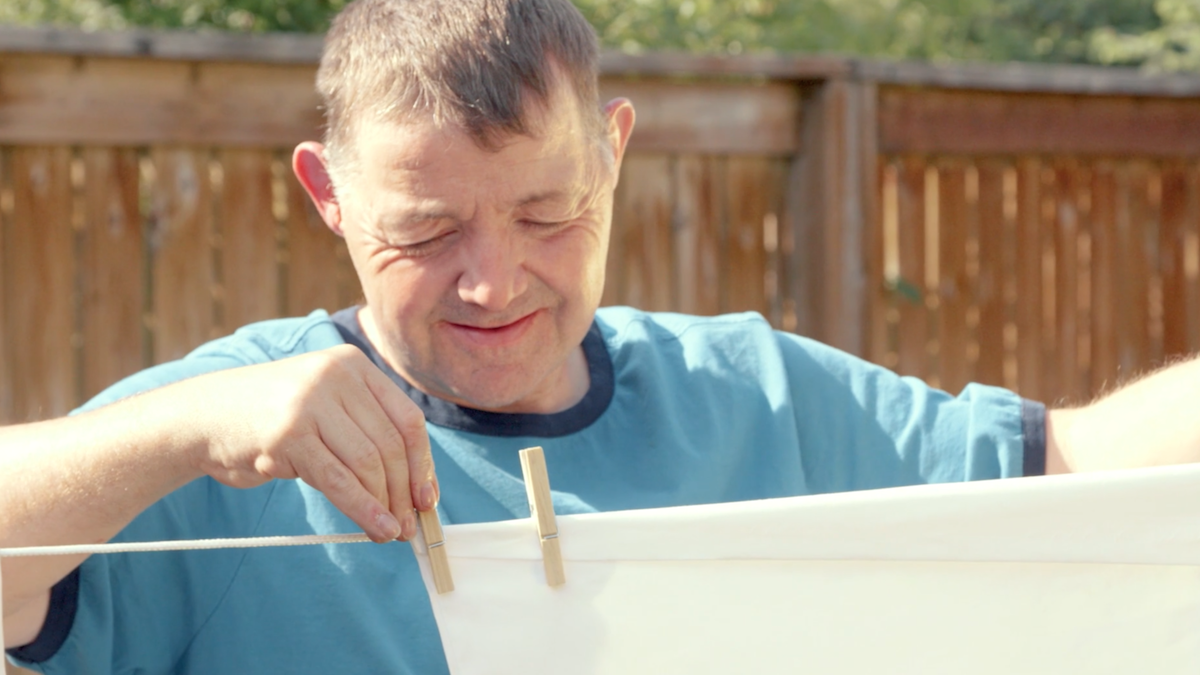 A man with a disability hanging up clothes on a clothes line outside in front of a wooden fence