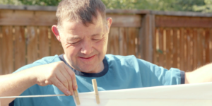 A man with a disability hanging up clothes on a clothes line outside in front of a wooden fence