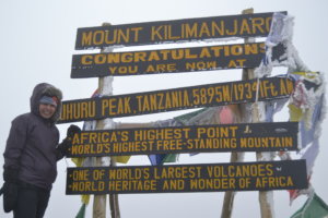 Sneha standing at the top of Mount Kilimanjaro with multiple signs behind her