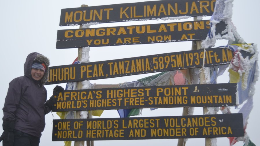 Sneha standing at the top of Mount Kilimanjaro with multiple signs behind her