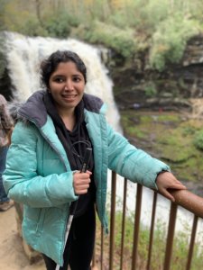 Baksha Ali smiling holding a white cane holding onto a railing next to a river with a waterfall behind her
