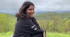 Baksha Ali smiling holding her white cane with a large forest in the distance behind her