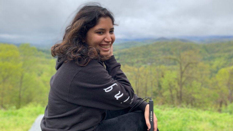 Baksha Ali smiling holding her white cane with a large forest in the distance behind her