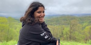 Baksha Ali smiling holding her white cane with a large forest in the distance behind her