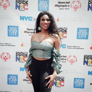 Lachi on a red carpet in front of logos for Disability Pride NYC, the Christopher and Dan Reeve Foundation, Special Olympics New York, and New York Life