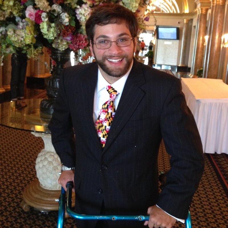 Aaron Kaufman smiling in front of flowers on a table, holding onto his walking frame