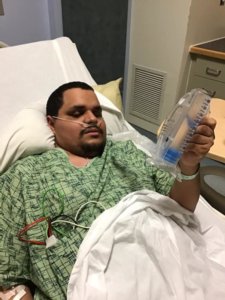 Leo Cantos lying in a hospital bed holding a tool for breathing exercises