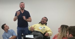 A man using a wheelchair next to an ASL interpreter speaks as three other people look on seated.