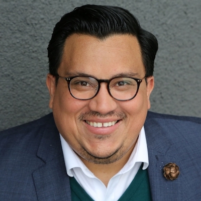 Jose Luiz Plaza smiling headshot. Plaza has black hair, is wearing glasses and a blue suit, and has a mustache.