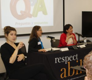 Debbie Fink smiles seated next to Carol Robles-Roman at a panel for Latinas with disabilities. Q&A sign behind them, ASL interpreter to the left.