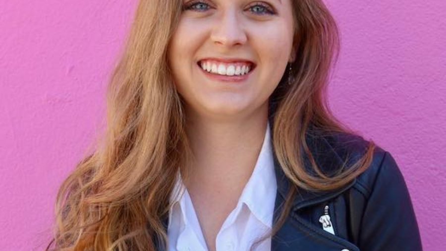 April Caputi smiling headshot. April is a white woman with long brown hair standing in front of a pink wall.