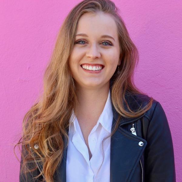 April Caputi smiling headshot. April is a white woman with long brown hair standing in front of a pink wall.