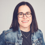 Carolyn Lertzman smiling headshot. Lertzman is a white woman with black hair down to her shoulders, wearing glasses, a necklace and a denim jacket.