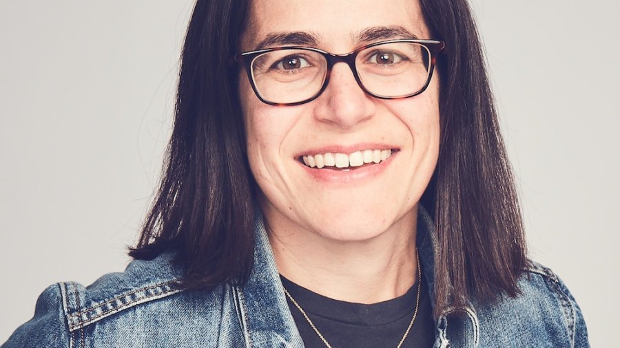 Carolyn Lertzman smiling headshot. Lertzman is a white woman with black hair down to her shoulders, wearing glasses, a necklace and a denim jacket.