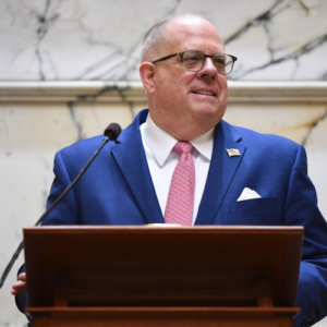 Governor Larry Hogan smiling, speaking behind a podium. Hogan is wearing a blue suit, red tie, and glasses