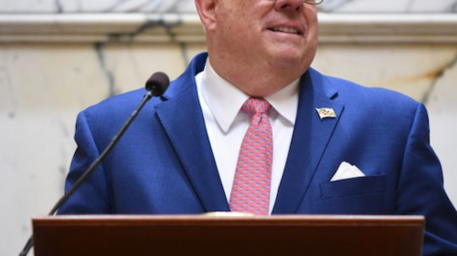Governor Larry Hogan smiling, speaking behind a podium. Hogan is wearing a blue suit, red tie, and glasses