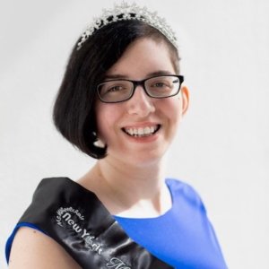 Lauren Arena headshot smiling wearing a sash that says Ms. Wheelchair New York on it. Arena is a white woman with black hair down one side of her head