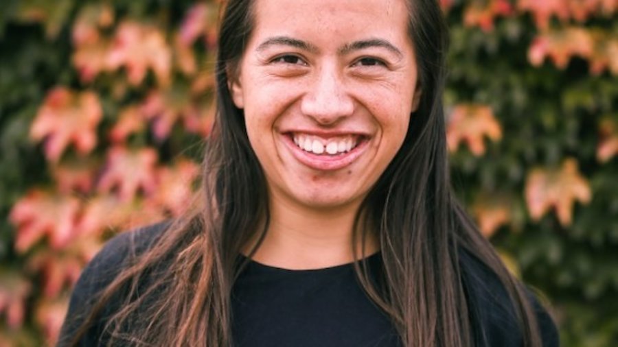 Nicole Homerin smiling headshot in front of bushes and flowers. Homerin has long brown hair and is wearing a black shirt