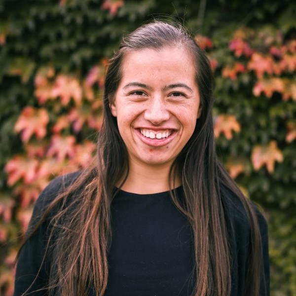 Nicole Homerin smiling headshot in front of bushes and flowers. Homerin has long brown hair and is wearing a black shirt