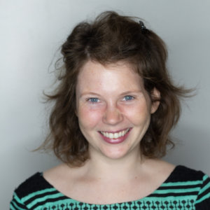 Sasha Ross Becker smiling in front of a white backdrop. Becker is white, has brown hair, and is wearing a black and green shirt