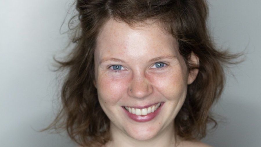 Sasha Ross Becker smiling in front of a white backdrop. Becker is white, has brown hair, and is wearing a black and green shirt