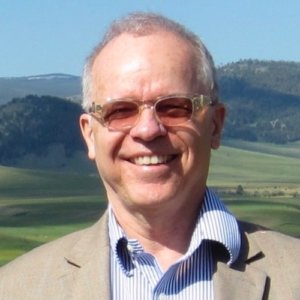 Timothy Gray smiling in front of grass and mountains wearing a suit and glasses. Tim is a white man with short white hair.