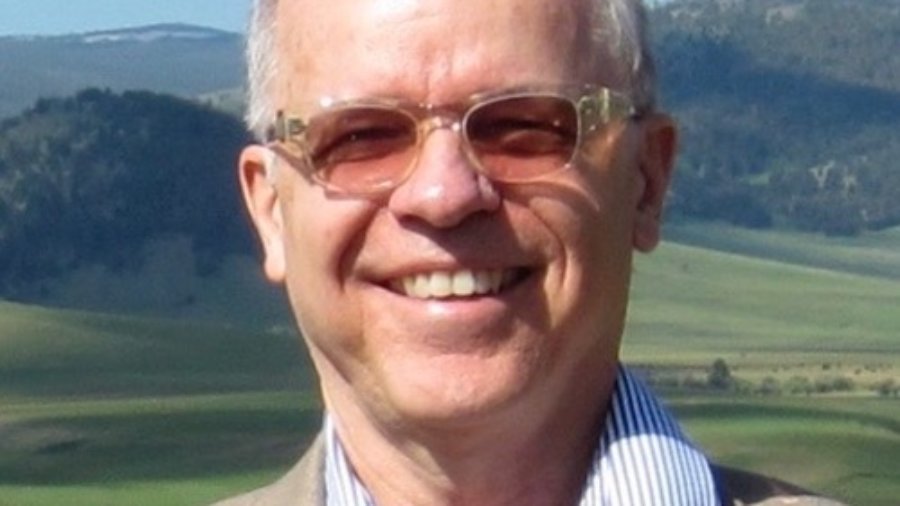 Timothy Gray smiling in front of grass and mountains wearing a suit and glasses. Tim is a white man with short white hair.