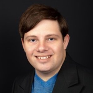 Zack Gottsagen smiling headshot wearing a black suit and blue shirt. Zack is a white man and has short brown hair.