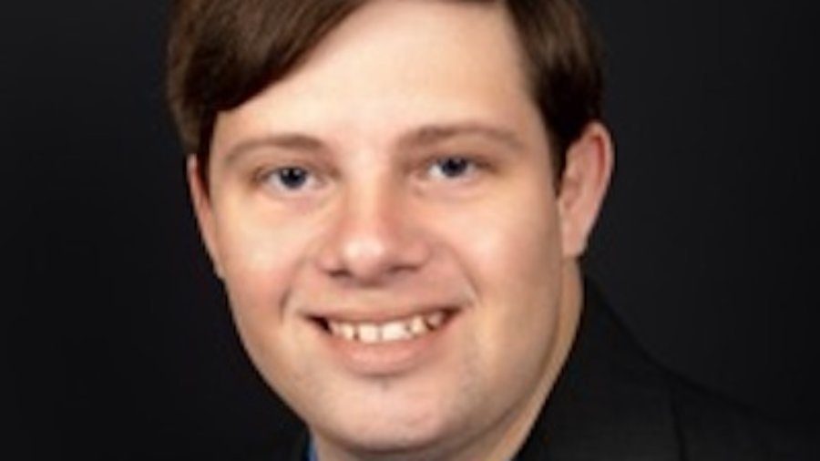 Zack Gottsagen smiling headshot wearing a black suit and blue shirt. Zack is a white man and has short brown hair.
