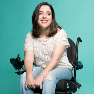 Anastasia Somoza smiling. Somoza is a white woman with brown hair down to her shoulders who uses a power wheelchair. She is seated in front of a green backdrop