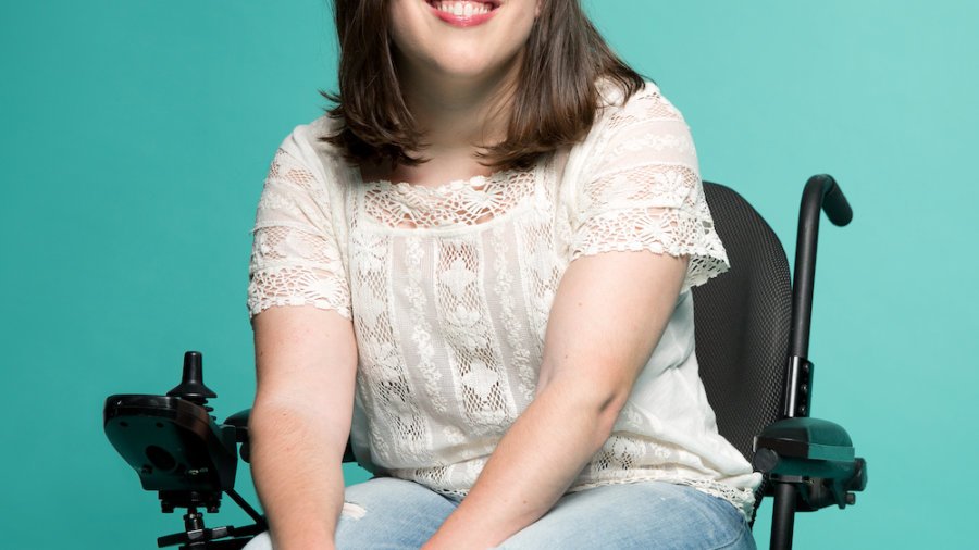 Anastasia Somoza smiling. Somoza is a white woman with brown hair down to her shoulders who uses a power wheelchair. She is seated in front of a green backdrop