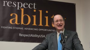 Brad Sherman speaking at the microphone with a RespectAbility banner behind him