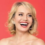 Eileen Grubba headshot smiling in front of a red backdrop. Grubba is a white woman with short blonde hair