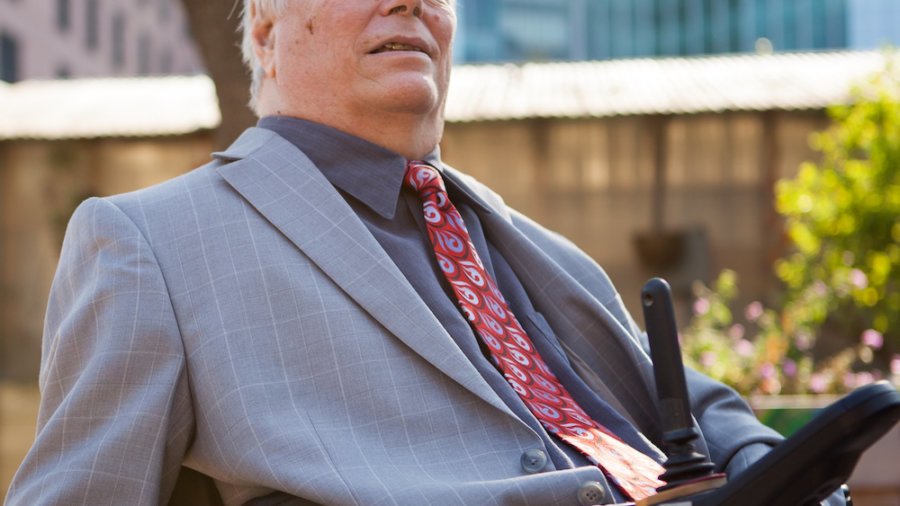 Lex Frieden smiling wearing a suit and tie. Frieden uses a power wheelchair.