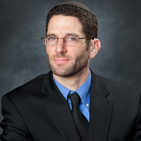 Rabbi Darby Leigh wearing a dark suit and tie smiling and looking at the camera against a charcoal photographers background