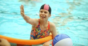 A young girl with a disability in a pool with one hand on the side of the pool and one arm up in the air.