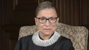 Justice Ruth Bader Ginsburg smiling wearing her robe, seated on a chair