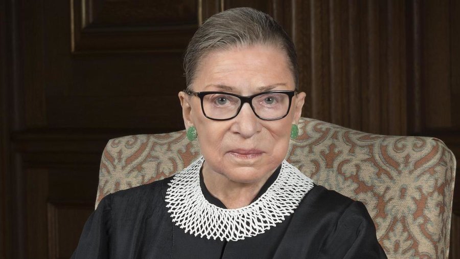 Justice Ruth Bader Ginsburg smiling wearing her robe, seated on a chair