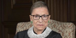 Justice Ruth Bader Ginsburg smiling wearing her robe, seated on a chair