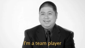 A man with a disability wearing a suit and tie speaking to camera in front of a white background. Caption: I'm a team player